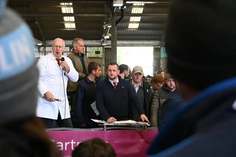 A man in a white coat and glasses speaks into a microphone at a gathering in a covered outdoor area, with several people standing behind him and in front of a pink banner.
