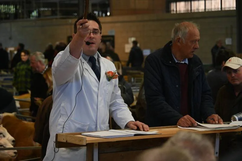 A man in a white coat with a microphone standing at a wooden table, surrounded by people and animals, in a busy indoor setting that appears to be a livestock or agricultural event.