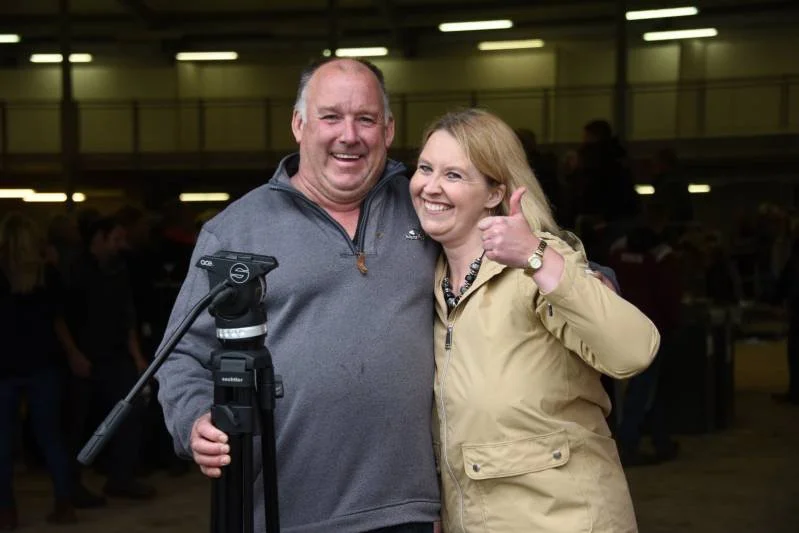 A man and woman smiling and posing for a photo indoors, with the woman giving a thumbs up. The man is holding a camera on a tripod.