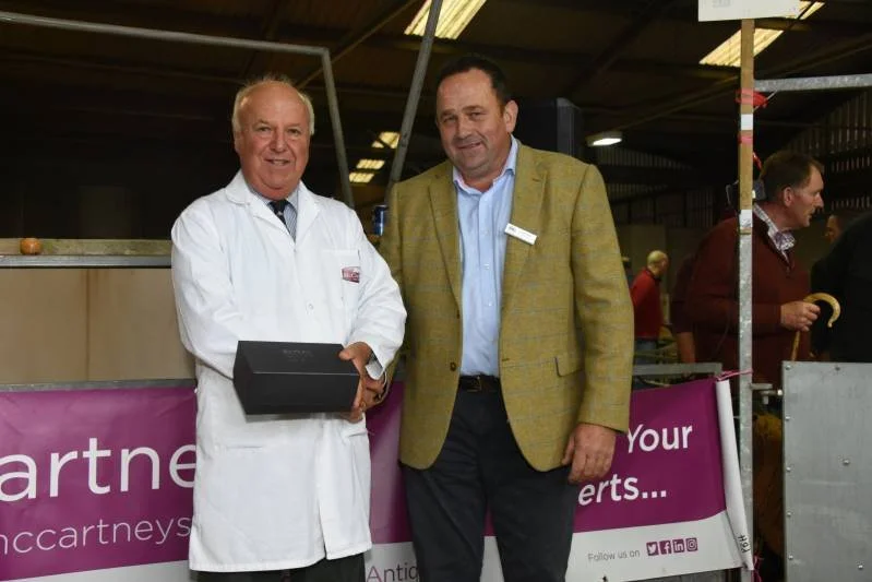 Two men standing together at an indoor event, smiling at the camera. The man on the left is wearing a white lab coat and holding a black box. The man on the right is dressed in a yellow blazer over a light blue shirt. Behind them is a purple banner w