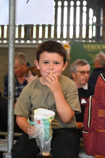 A young boy with short brown hair and a khaki t-shirt sitting on a bench, holding a snack in one hand and a bag of chips in his lap, smiling at the camera.