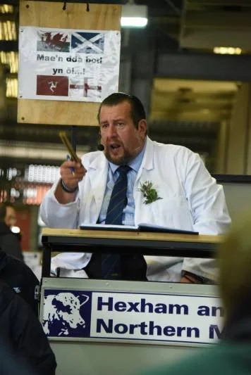 Man in white lab coat holding a pen, speaking at a desk with a sign that reads 'Hexham and Northern M' in an industrial setting.