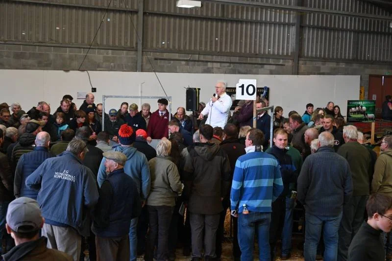 A large indoor gathering with many people listening to a man speaking into a microphone on a stage, with a sign displaying the number 10 behind him.