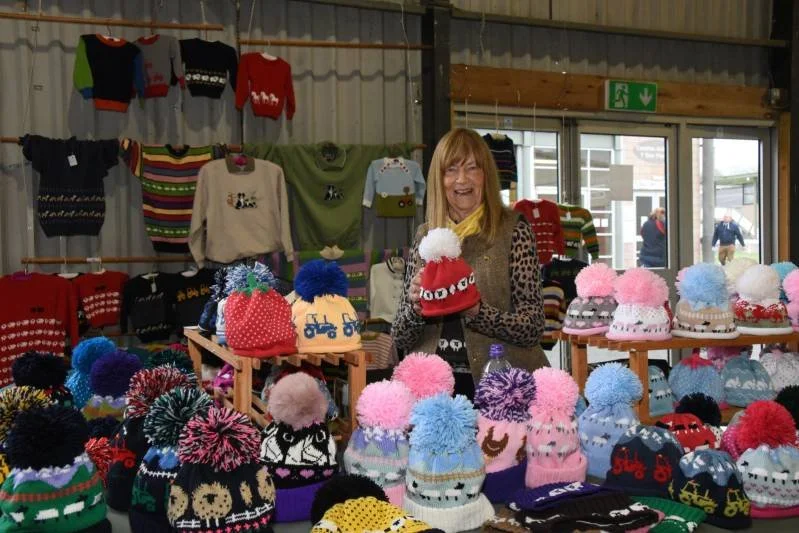 A woman shopping at a holiday knit hat booth, with shelves of colorful hats and sweaters, inside a retail store.