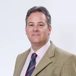 Professional man with short grey hair wearing a beige blazer, light pink shirt, and dark patterned tie, smiling at the camera