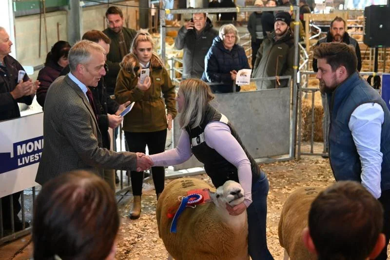 A woman shaking hands with a man at an agricultural event, with sheep nearby and several onlookers observing.