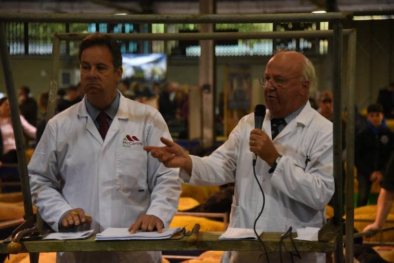 Two men in white coats speaking at a public event, one holding a microphone and gesturing, with a crowd in the background.