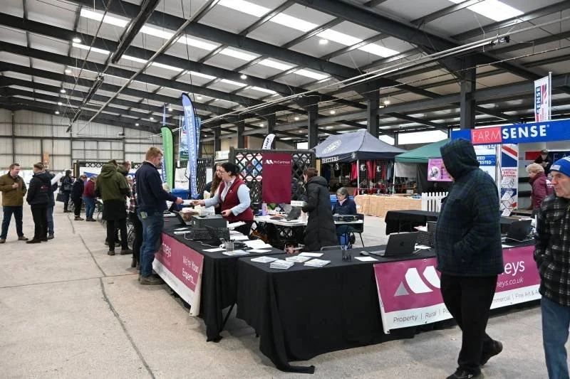 People attend an indoor expo with vendor booths, some checking in at tables with black tablecloths, while others browse stalls selling various products and services.