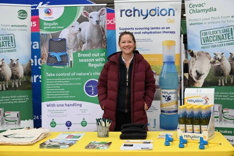 A woman standing behind a table at an agricultural or veterinary exhibition booth. The booth displays various promotional banners for livestock health products, including rehydion Gel and Cevac Chlamydia. The table is covered with informational mater