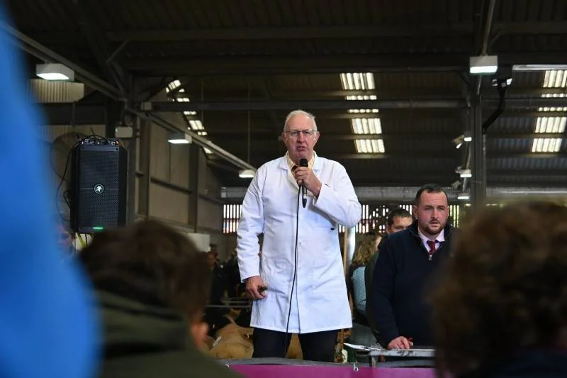 An older man in a white coat speaking into a microphone at a gathering inside a large industrial-style building.