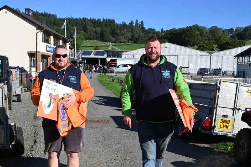Two men walking outdoors near a racecourse or event venue, each holding a box and wearing casual clothing with one in an orange jacket and the other in a green and navy hoodie, with several trailers and buildings in the background.