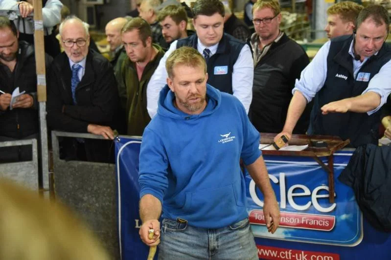 A man with a beard and a blue hoodie demonstrating a product in front of a group of people at an indoor event, with several people watching attentively.