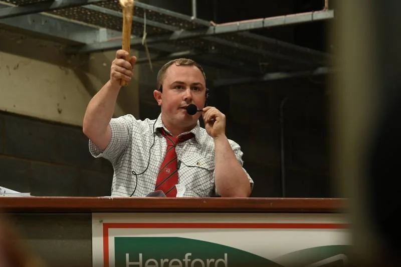 A man with a microphone in his hand and a red tie is speaking at a rally or event, raising his arm. There is a sign with the word "Hereford" partially visible in front of him.