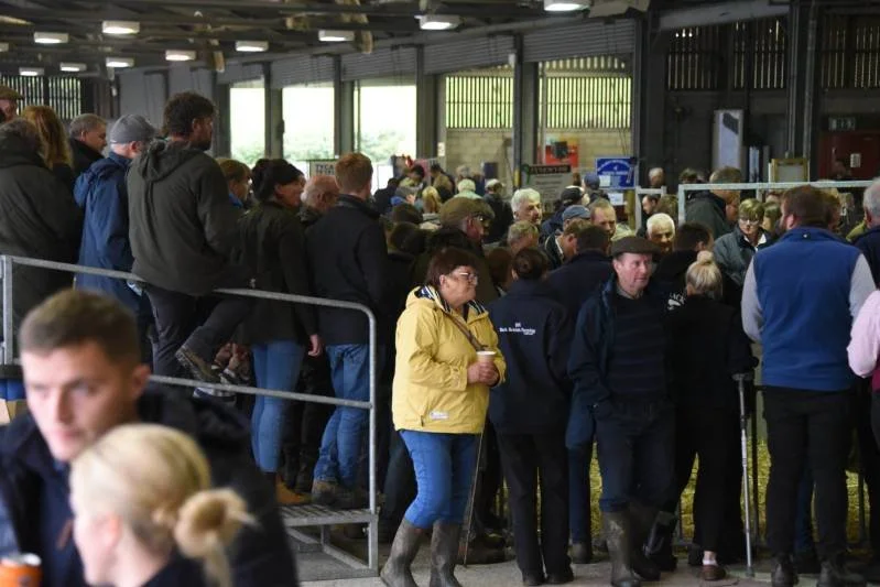 Crowd of people standing in line indoors, with some holding cups, in a large industrial-style building with large windows.