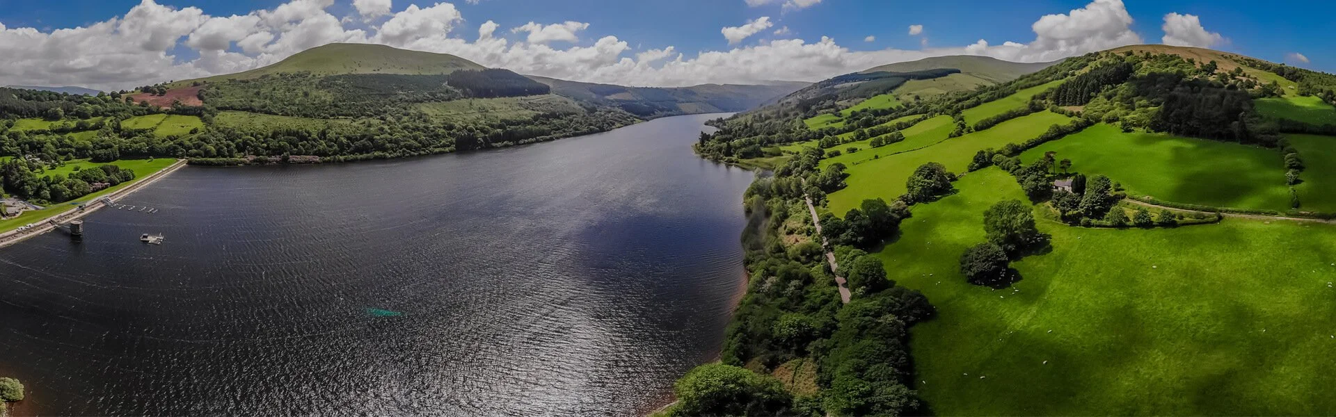 Aerial view of a large river or lake surrounded by green hills and forests under a partly cloudy sky.