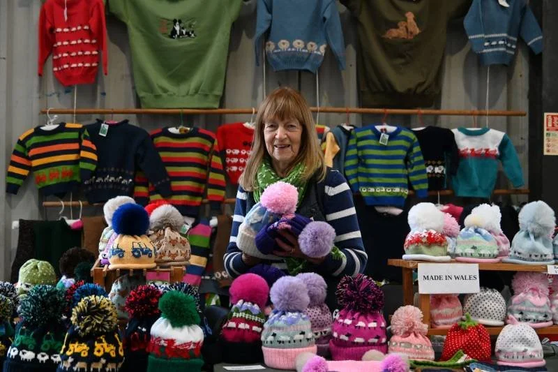 A woman selling colorful knitted hats and sweaters at a craft fair, with various winter clothing items displayed behind her.