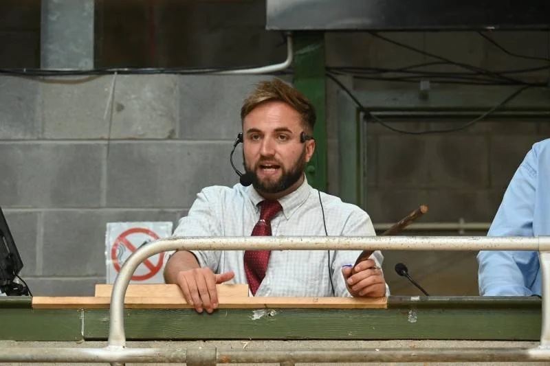 Man with a beard wearing a white shirt, red tie, and headset microphone speaking at a podium with a green railing, in an industrial-style setting.