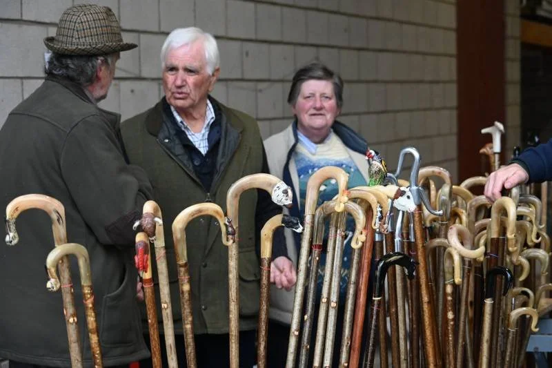 Three elderly people looking at walking canes displayed on a table at an indoor market or shop.