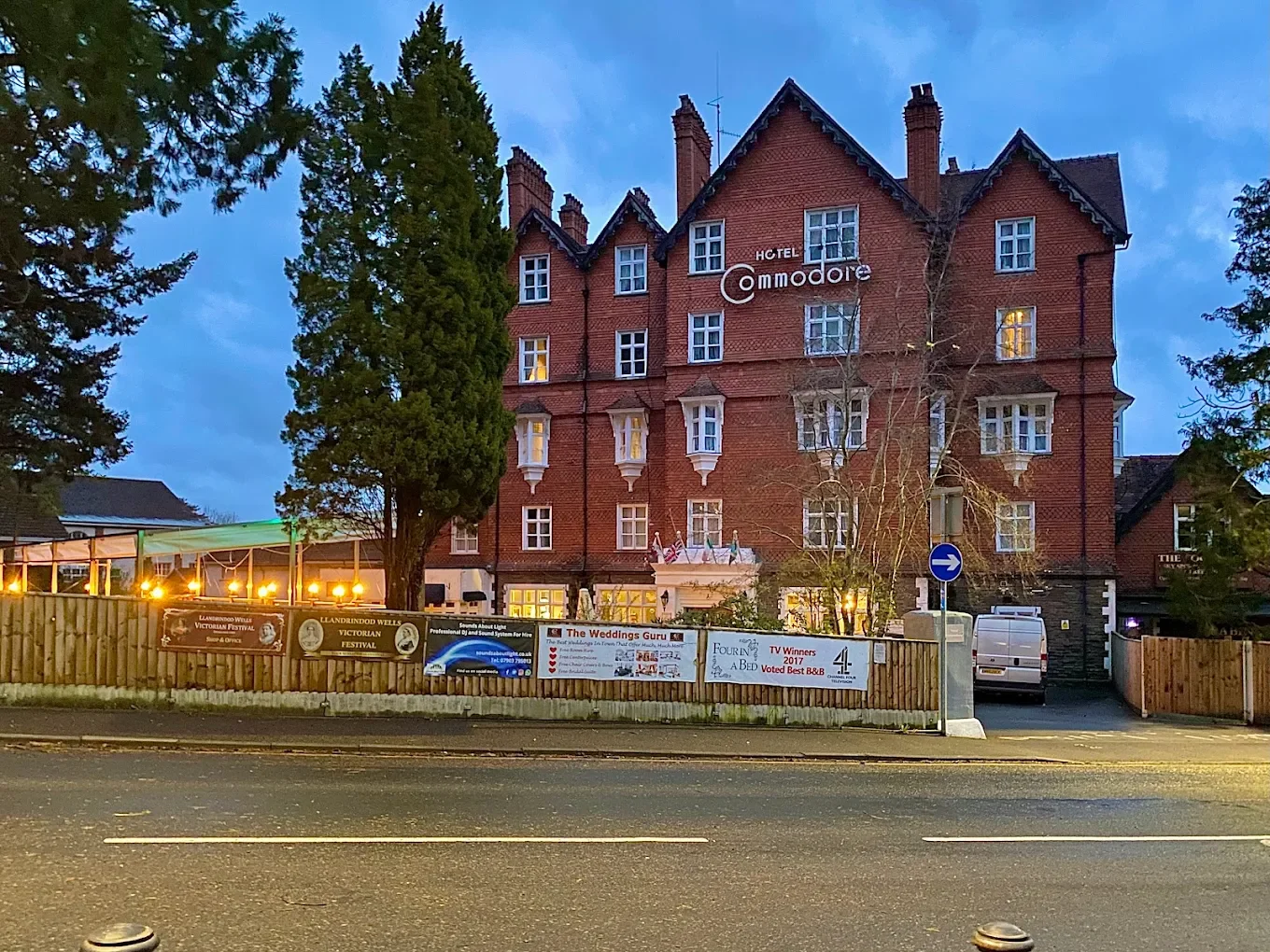 A large red brick hotel named Hotel Commodore at dusk with trees in front and a street with parked cars and signs.