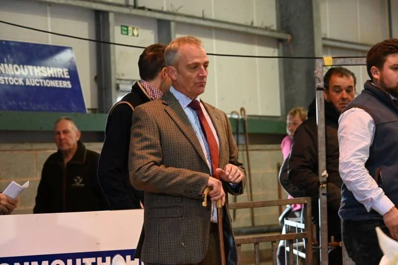 A group of people standing in line at an indoor event, with a man in the foreground wearing a brown checkered blazer and red tie, holding a ticket or card, and other individuals behind him.
