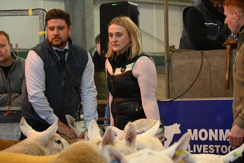 People observing livestock at an agricultural event, including a woman and a man in focus, with sheep in the foreground.