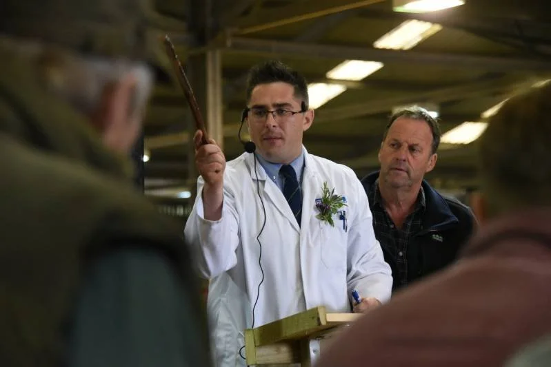 A man in a white lab coat and glasses is holding a pointer and speaking to a group of people in an industrial setting. Another man is standing nearby, listening. The group appears to be engaged in a presentation or discussion.
