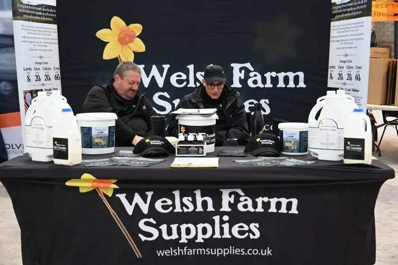 Two men sitting at a table with Welsh Farm Supplies branding, displaying pet products and farm supplies at an indoor event.