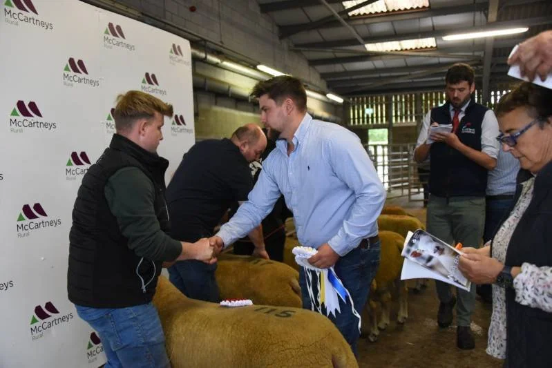 Men and women at a livestock event shaking hands and examining sheep inside a barn, with a McCartney's branding backdrop.
