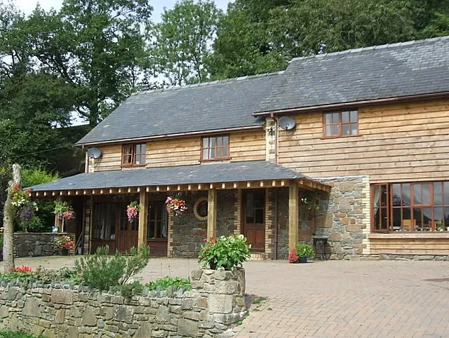 A two-story house with a wooden exterior, stone accents, hanging flower baskets, and a brick driveway with a stone wall in front.