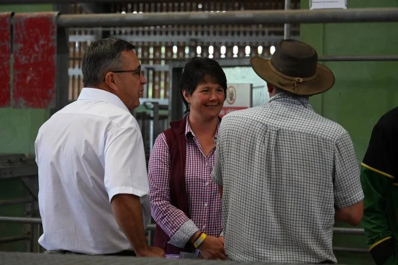 Three people engaged in conversation: a man with glasses in a white shirt, a woman with short dark hair in a red and white checkered shirt with a vest, and a man in a checkered shirt and large hat, in an outdoor setting with a green wall and metal ra