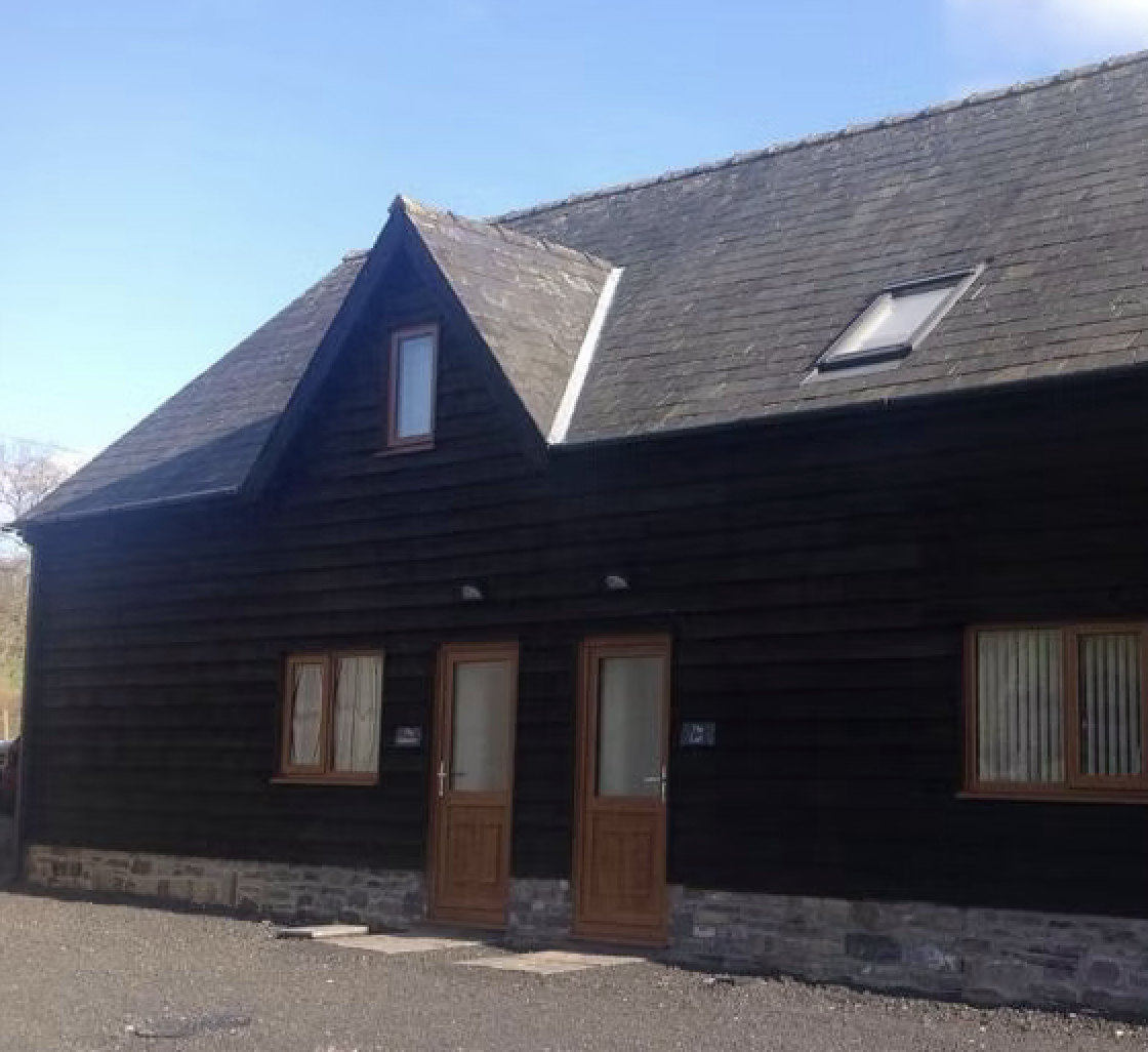 A black wooden house with a stone foundation, featuring two wooden doors and three windows, one on the upper floor and two on the ground level, under a clear blue sky.