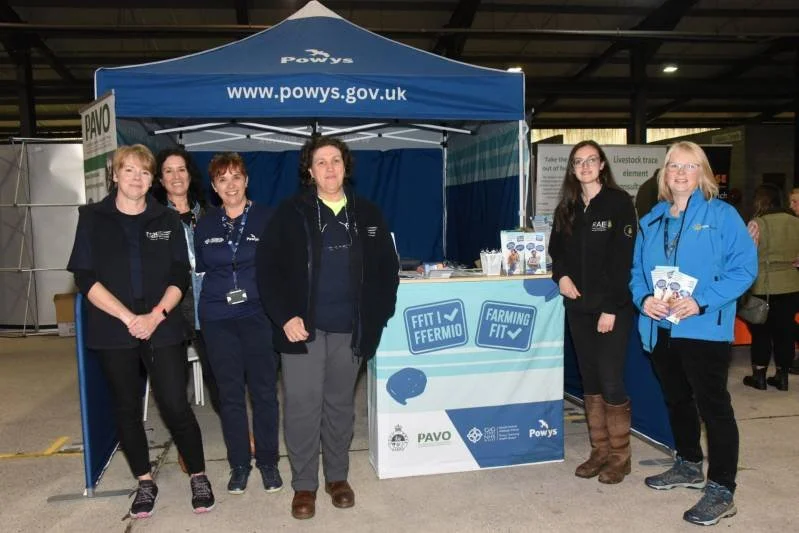 Five women standing in front of a booth with a blue canopy at an indoor event. The booth displays the website www.powys.gov.uk and has signs promoting FIT and FARMING FIT. The women are smiling and dressed in casual and outdoor clothing, some wearing