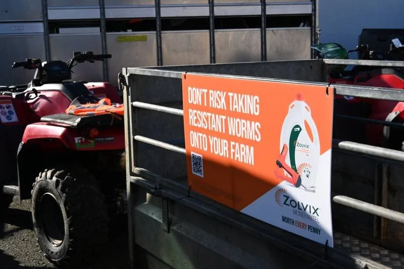 Sign on a farm trailer warning about resistant worms, with a QR code and pesticide logo, parked next to red and black all-terrain vehicles.