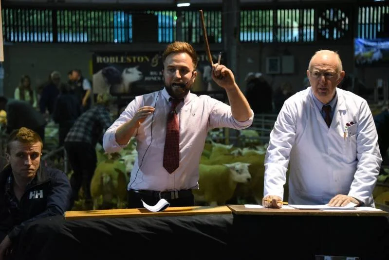 Two men standing at a table, one gesturing passionately, the other looking serious, with a livestock auction in the background.