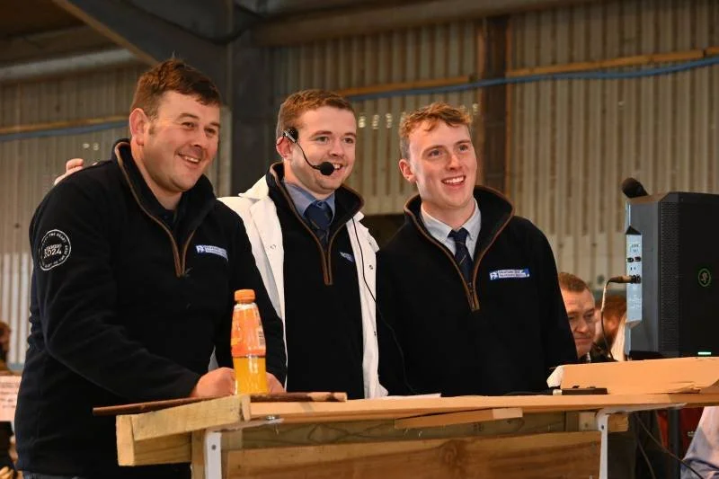 Three young men standing behind a wooden desk in an indoor setting, smiling and watching a computer screen, with a bottle of orange drink on the desk. The man in the middle is wearing a headset and a white jacket, possibly indicating he is working or