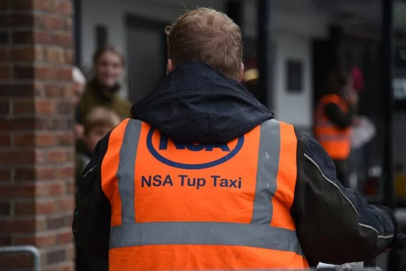Person wearing an orange reflective vest with the text 'NSA Tup Taxi' and the NSA logo, standing outdoors with people in the background.