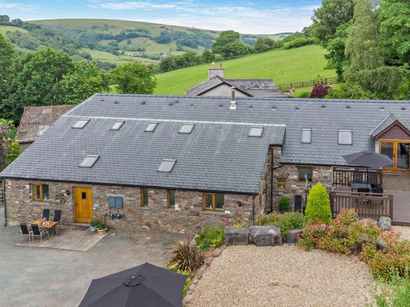 Stone house with gray slate roof and yellow door, outdoor dining area with chairs and table, garden with plants and rocks, surrounding lush green hills and trees.