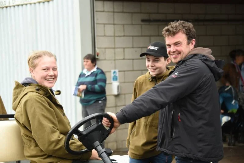 A group of people smiling and interacting, with a man holding a steering wheel and a woman sitting nearby in an outdoor setting near a concrete building.
