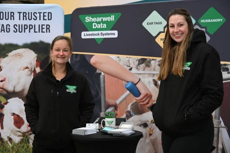 Two women standing behind a booth at an event, one smiling and the other with a neutral expression, with a banner in the background displaying 'Shearwell Data' and 'Livestock Systems,' featuring images of cows and information about ear tags and husba