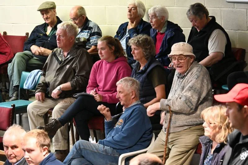 Group of older adults and a young woman sitting in a room, some engaging in conversation, wearing casual clothing.