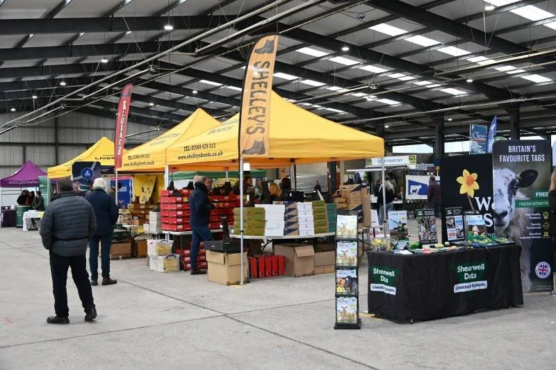 Indoor market or trade fair with yellow and purple tents, people browsing stalls with boxes and products, and informational stands about Shearwell Data and animal tags.