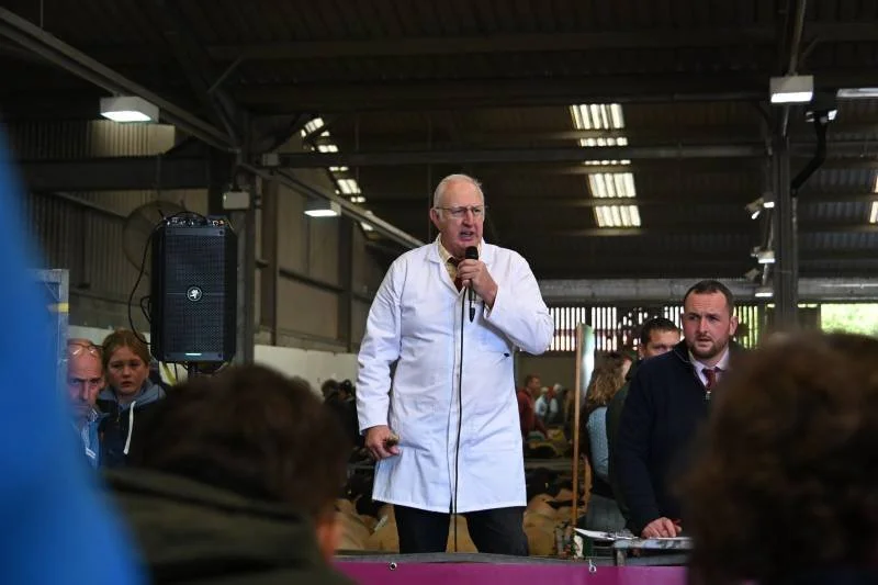 A man in a white lab coat speaking into a microphone at a gathering or event, with people sitting and listening in an industrial-style setting.