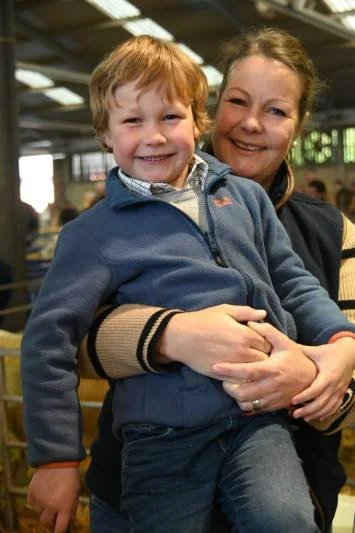 A young boy being held and hugged by an adult woman, both smiling, in an indoor setting with metal beams and natural light.