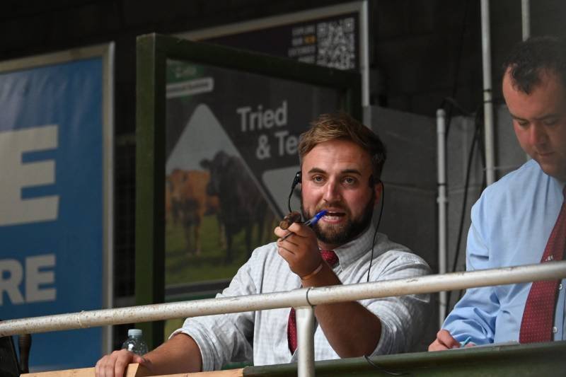 A man with a headset and a microphone speaking from a broadcast booth, with another man standing next to him in a blue shirt.