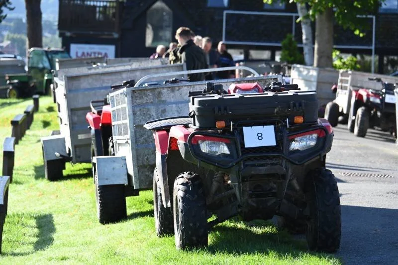 A red utility vehicle with a black front rack, parked on grass, with several trailers and people in the background.