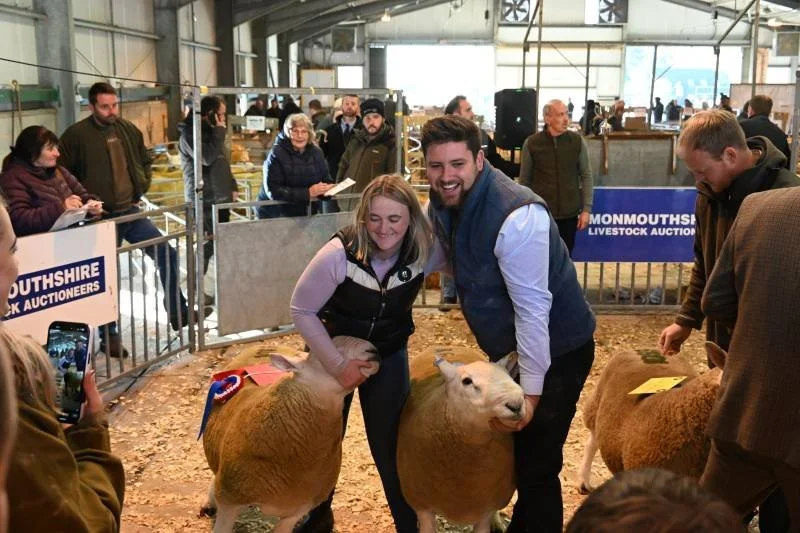 People at a livestock auction with sheep, some holding lambs, in an indoor barn setting.