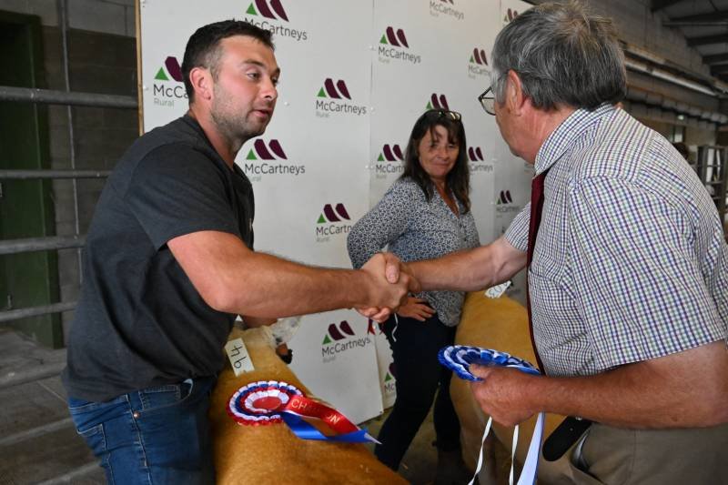 Two men shaking hands at a rural event, with one holding rosette ribbons. A woman stands behind them, and the background displays a McCartneys logo on a white backdrop.