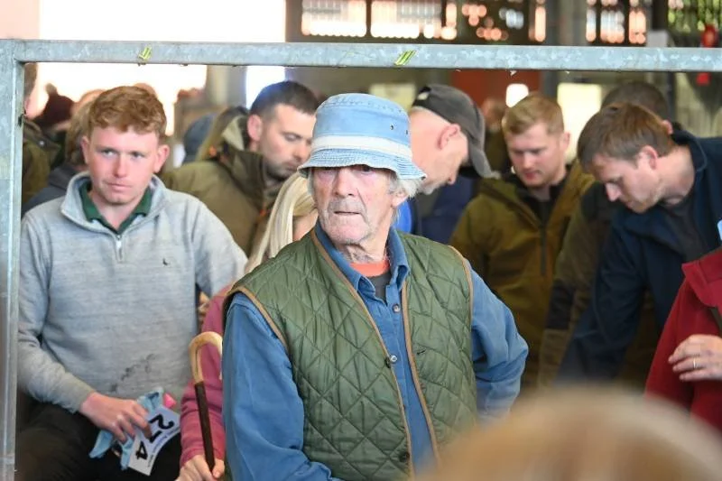 An elderly man in a blue bucket hat and green vest stands among a group of younger men indoors.