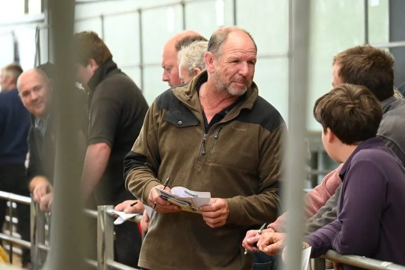 A man in a brown and black jacket holding a booklet, talking to a group of people in an indoor setting.