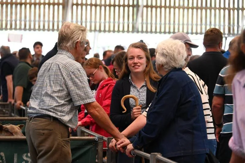 An elderly couple shaking hands with a young woman at a crowded indoor event, with other people and a green livestock pen in the background.